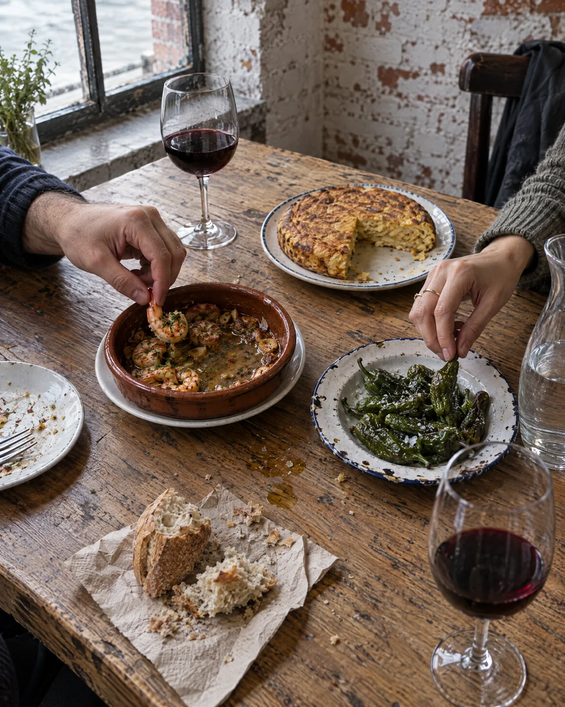 Ein Holztisch am Fenster mit geteilten Tapas: Gambas in der Cazuela, angeschnittene Tortilla, Pimientos de Padrón, Brot auf Papier, zwei Gläser Rotwein, zwei Hände greifen zu