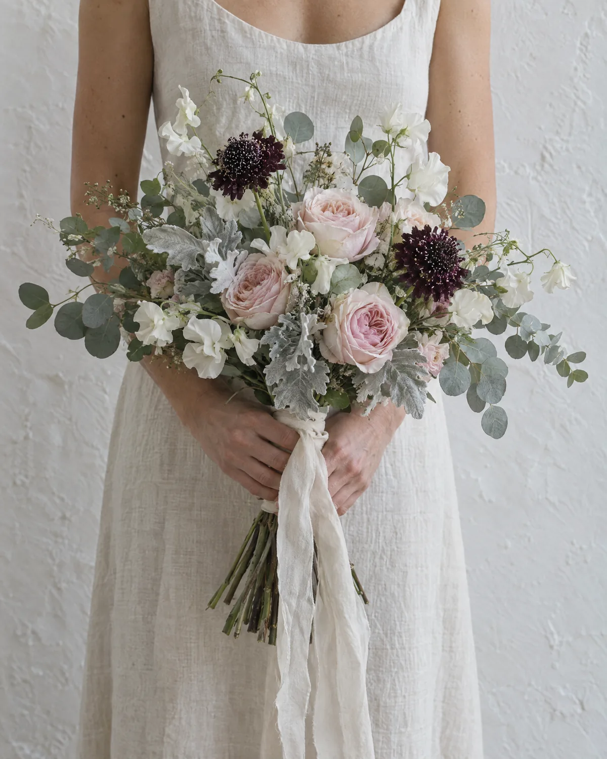 Lockerer Brautstrauß aus puderrosa Gartenrosen, cremefarbenen Wicken, Eukalyptus und zwei dunklen Scabiosen, in beiden Händen vor einem Leinenkleid gehalten, Seidenband hängt herab